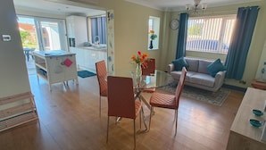 A living room with a glass dining table, chairs, a sofa, and a kitchen in the background.