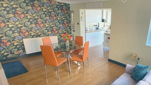 A dining area with a glass table and orange chairs, a patterned wallpaper, and a view into the kitchen.