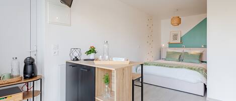 Een kleine kamer met een bed, een kitchenette, een televisie en een wandplank.