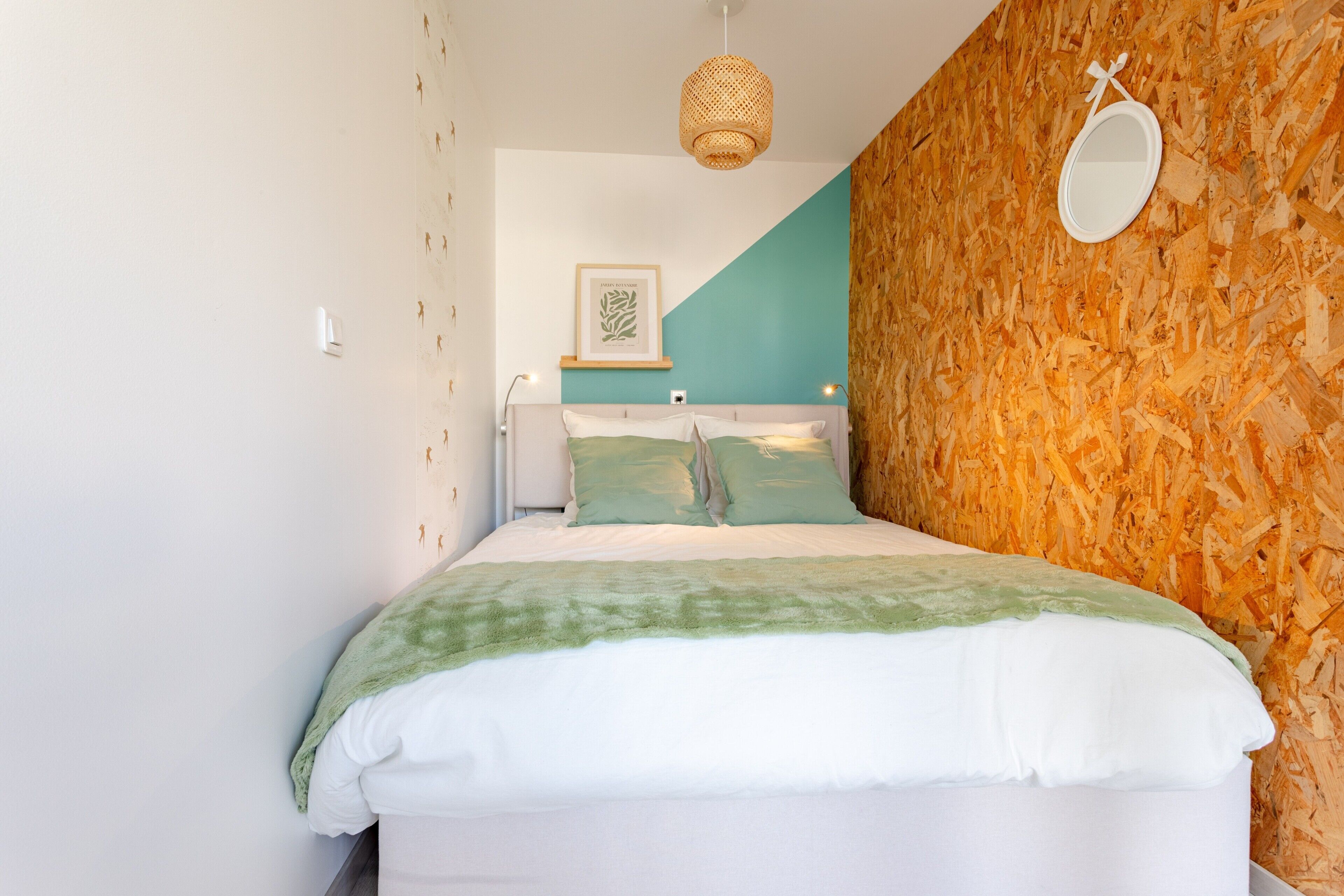 Una cama con colcha verde y blanca, un espejo montado en la pared y una obra de arte enmarcada sobre la cama.