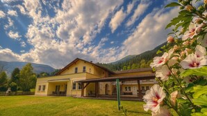 A two-story yellow building with a covered porch, surrounded by a grassy area and mountains in the background. - Appartementhaus Flattach (Flattach)