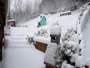 A snow-covered outdoor area with a wooden fence, a small blue shed, and a stone birdhouse.