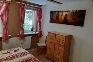 A bedroom with a bed, a wooden dresser, a chair, a window with curtains, and a forest-themed painting on the wall.