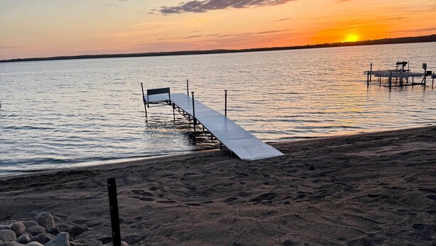 Ein Strand mit einem hölzernen Steg, der sich ins ruhige Wasser erstreckt, umgeben von Felsen und einem Sonnenuntergang im Hintergrund.