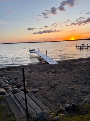 Una playa con un muelle de madera que se adentra en aguas tranquilas, rodeada de rocas y con un atardecer al fondo.