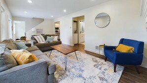 Ein modernes Wohnzimmer mit einer Couch, einem Sessel und einem Couchtisch. Im Hintergrund ist eine Küchenzeile erkennbar.