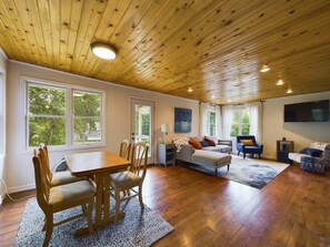 Un salón amplio con techo de madera, una zona de comedor con mesa y sillas de madera, un sofá y un televisor montado en la pared.