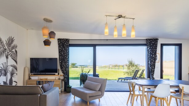 Ein modernes Wohnzimmer mit Flachbildfernseher, einer Couch, einem Esstisch mit Stühlen und einem großen Fenster mit Blick auf eine Rasenfläche.
