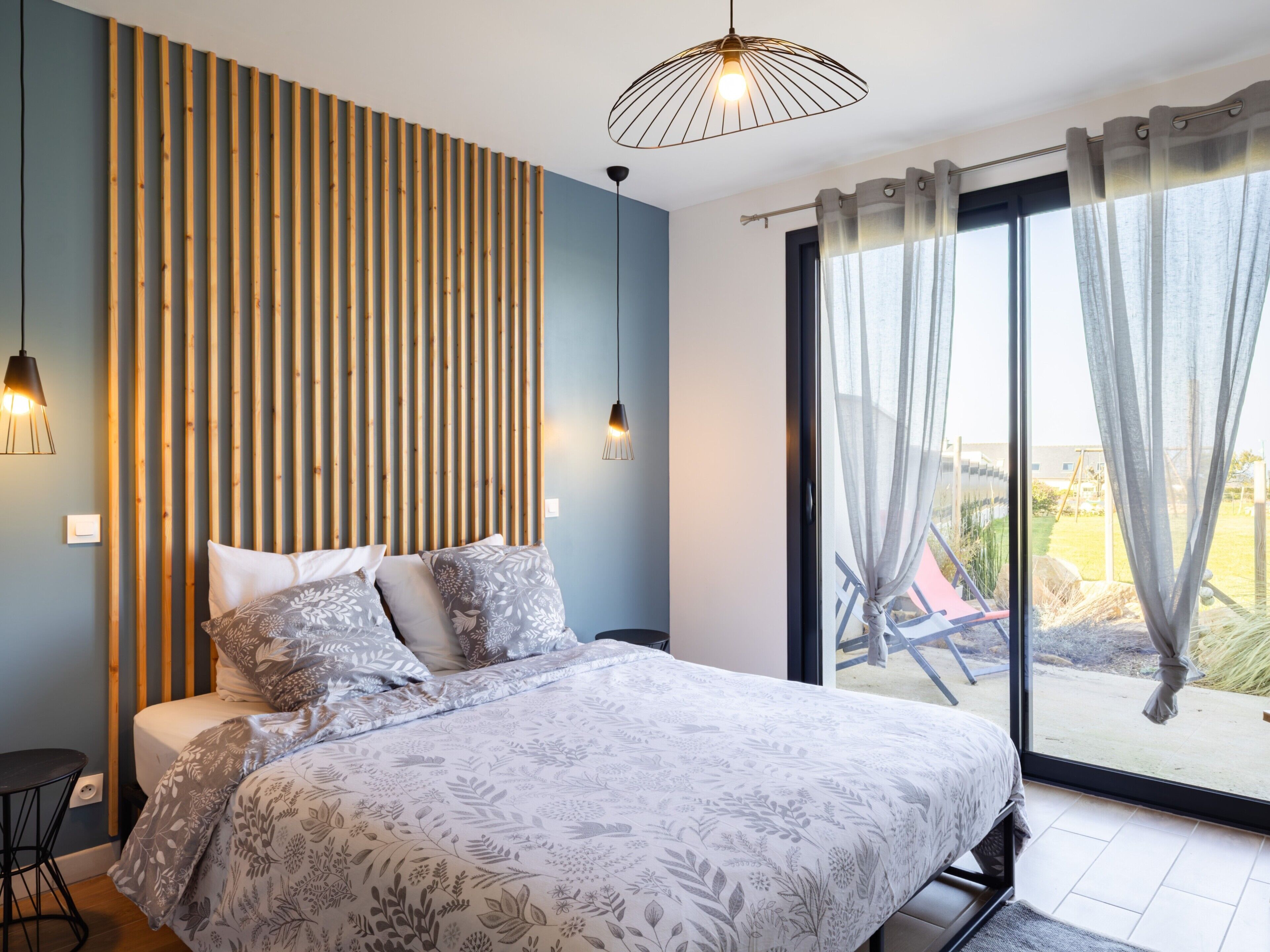 Une chambre avec un lit, un mur recouvert de lambris en bois, une grande fenêtre avec des rideaux et un luminaire suspendu.