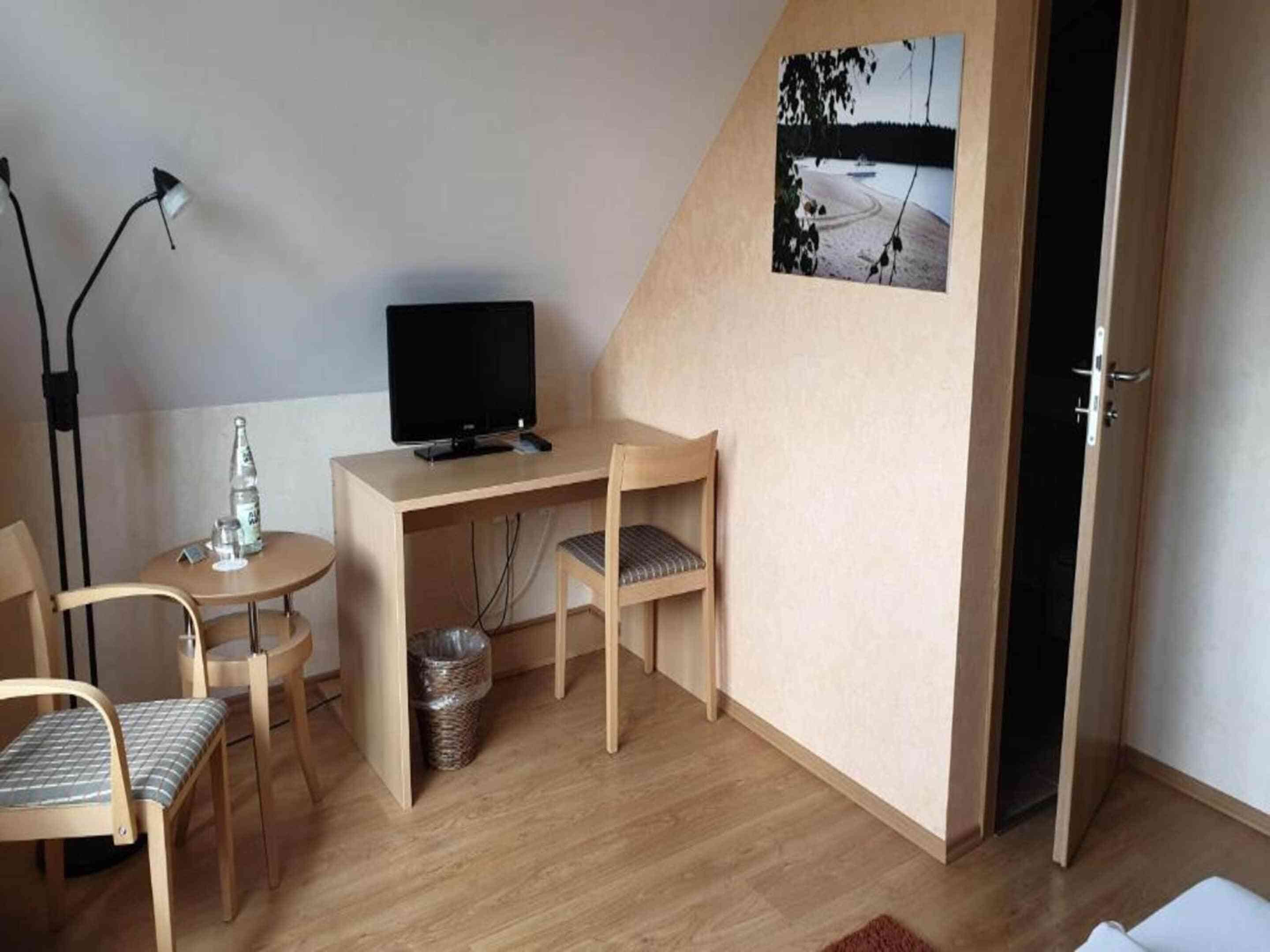 Quarto pequeno com uma escrivaninha, uma cadeira, uma televisão e um quadro na parede.