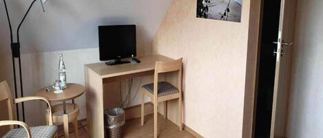 Quarto pequeno com uma escrivaninha, uma cadeira, uma televisão e um quadro na parede.