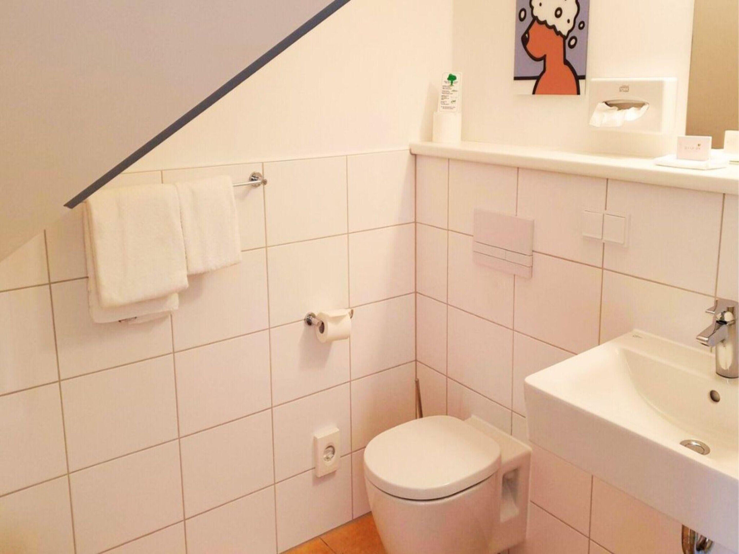 A small, clean bathroom with white tiles, a toilet, a sink, and a towel rack.