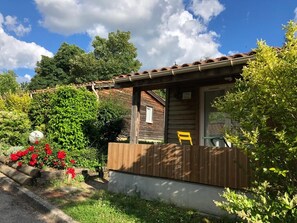 Eine kleine Holzhütte mit Veranda, umgeben von üppiger Vegetation und blauem Himmel.