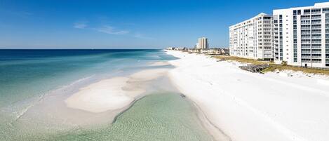 A beach with clear shallow waters, a sandy shore, and a row of white buildings with multiple floors and balconies.