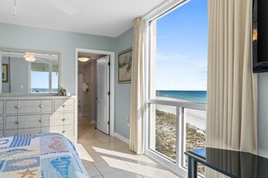 Ein Schlafzimmer mit großem Fenster und Blick auf den Strand, einem Bett mit blau-weiß gestreifter Bettdecke, einem Schminktisch und einer Tür zu einem Badezimmer.