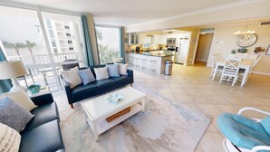 Een moderne woonkamer met een zwarte bank, een witte salontafel en uitzicht op de keuken en het balkon.