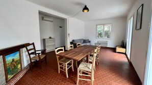 A dining area with a wooden table, chairs, a pendant light, a sofa, a TV stand, and a window. - Holiday Home “L'angèle - Spacious With” with Private Terrace, Wi-Fi & Air Conditioning (Portel-des-Corbières)