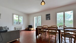 A modern living room with a sofa, TV stand, dining table with chairs, large windows, and a hanging light fixture. - Holiday Home “L'angèle - Spacious With” with Private Terrace, Wi-Fi & Air Conditioning (Portel-des-Corbières)
