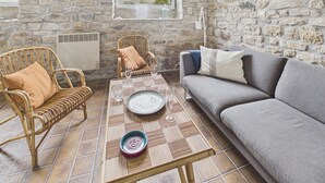 A cozy room with a stone wall, a grey sofa, wicker chairs, a tiled floor, and a window overlooking houses.