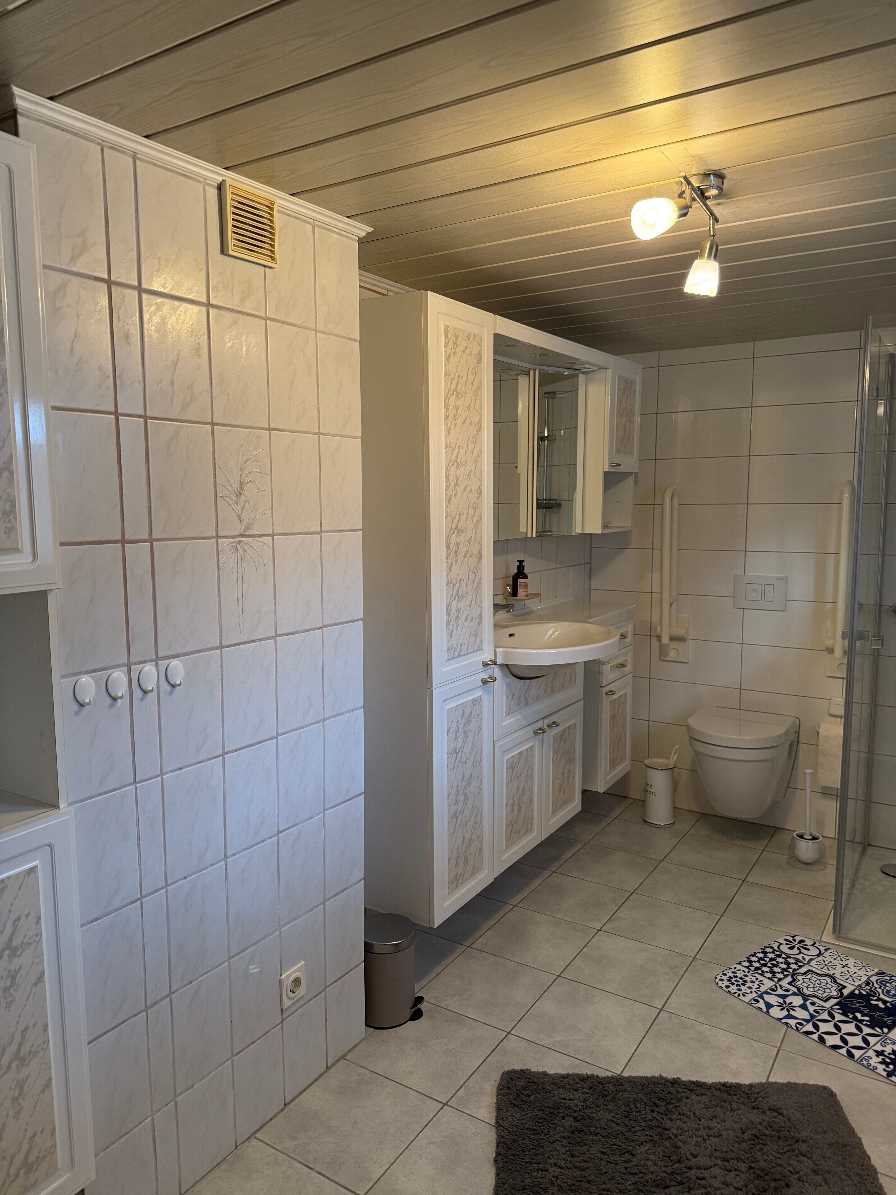 Ein Badezimmer mit weiß gefliesten Wänden, einem Waschbecken, einer Toilette, einer Dusche und einem kleinen Mülleimer.