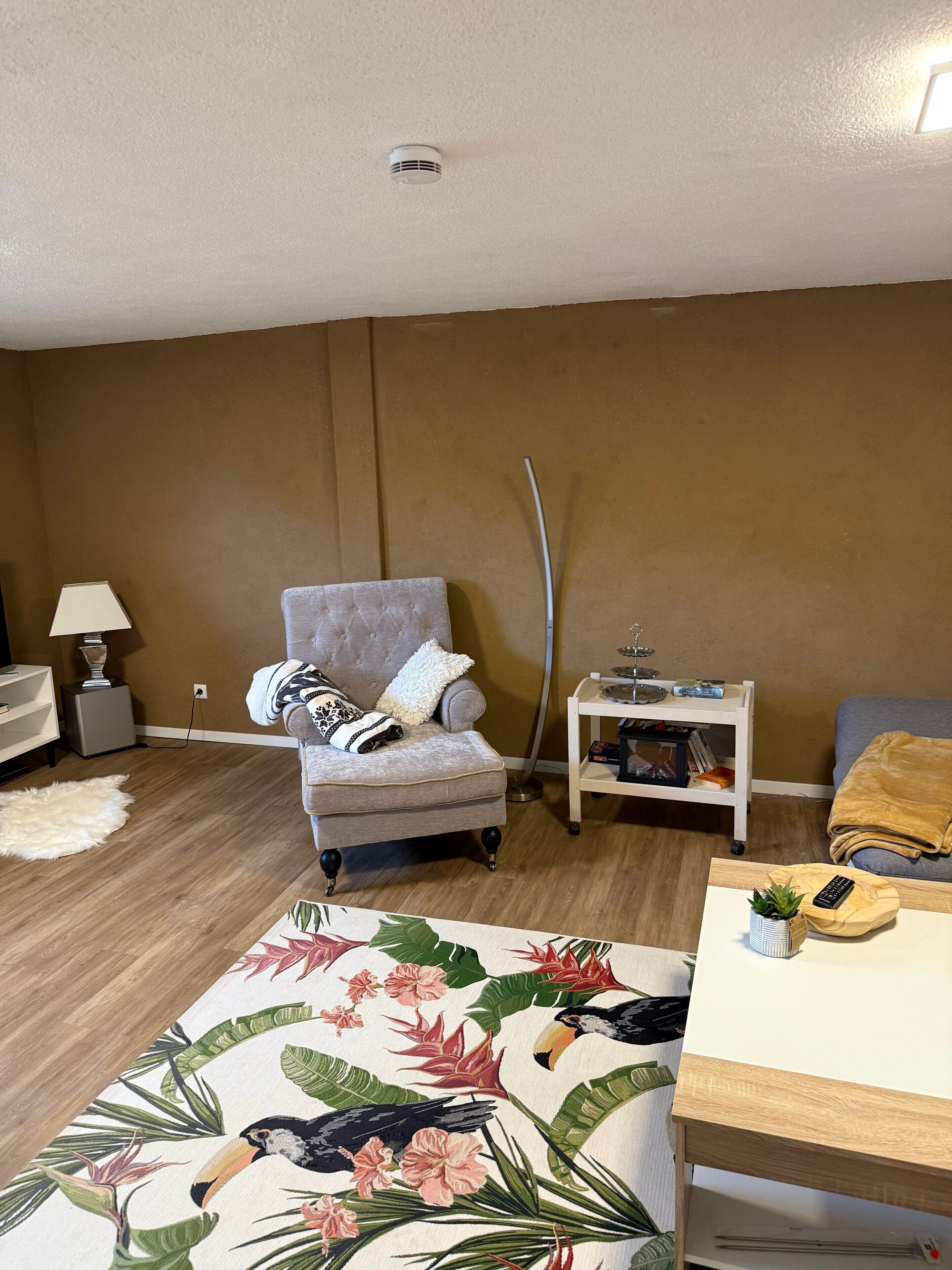 A living room with a grey armchair, a white side table, a small white lamp, a wooden coffee table, and a patterned rug.