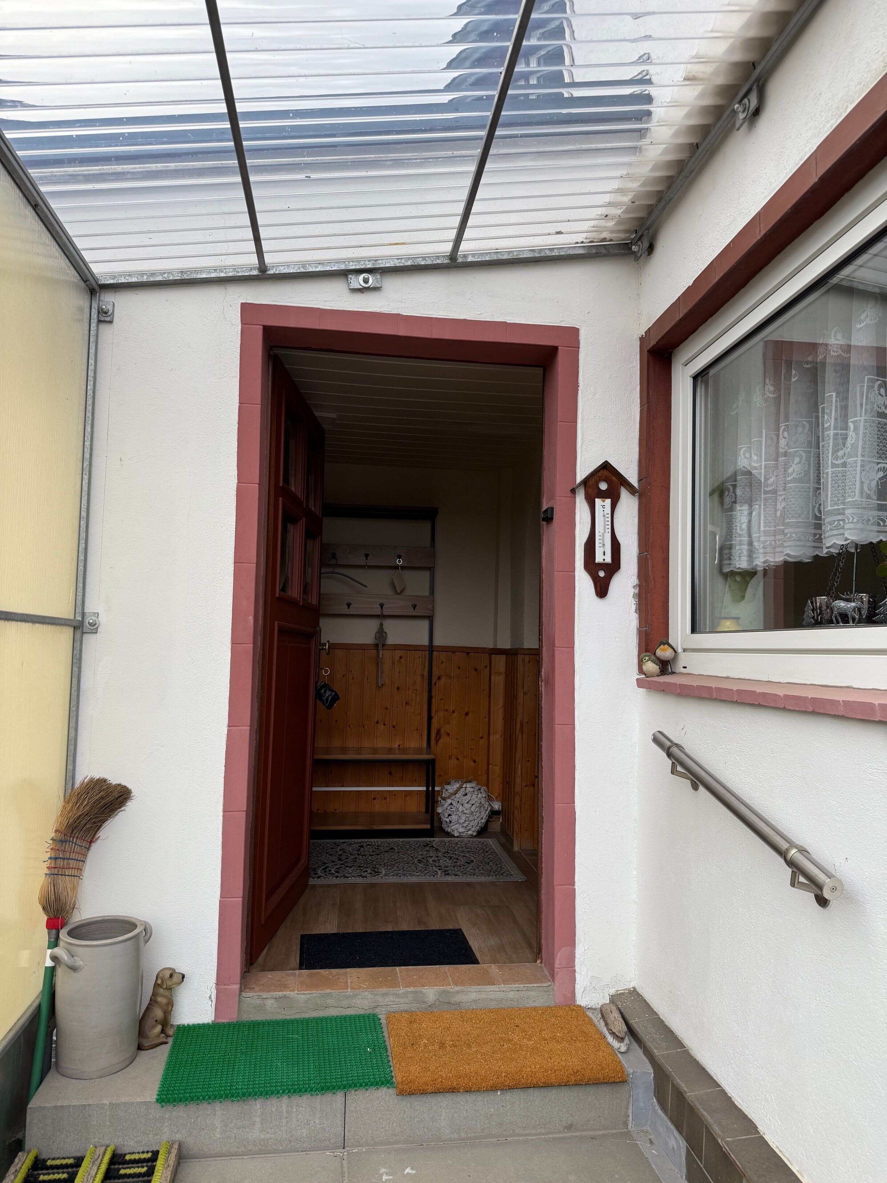Una entrada de casa con puerta de madera, un perro pequeño, un felpudo verde, un felpudo marrón y una ventana con adornos.
