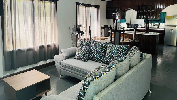 A living room with a sofa, a coffee table, and a fan. There is a kitchen area with a stove and cabinets.