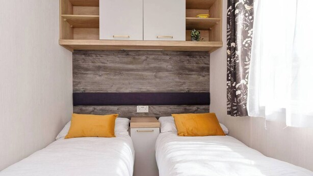 Une chambre avec deux lits simples, une tête de lit en bois, une petite étagère en bois et une fenêtre avec des rideaux.
