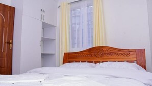 Une chambre à coucher avec une tête de lit en bois, du linge de lit blanc, une armoire et une fenêtre avec des rideaux.