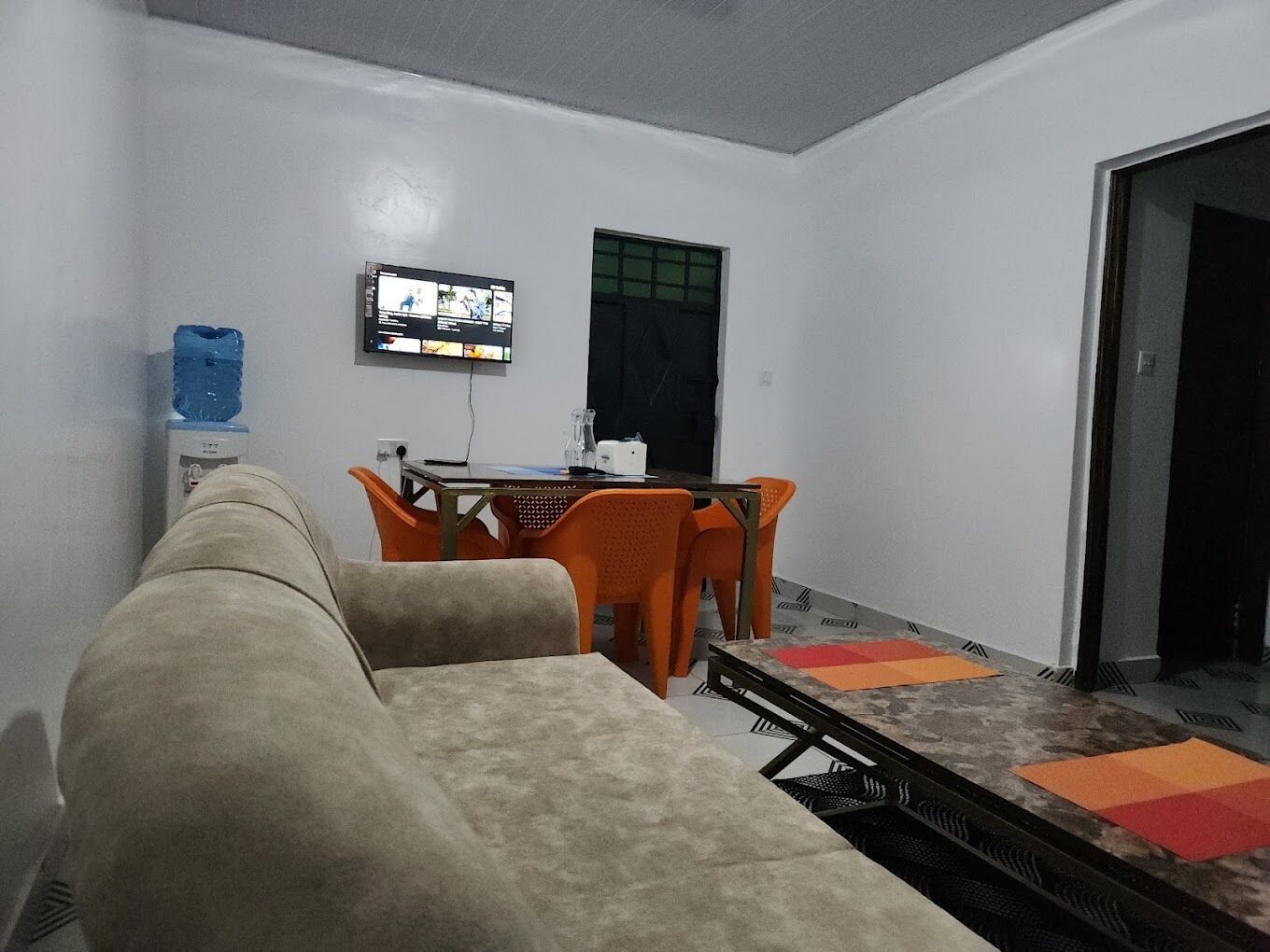 Una habitación pequeña y sencilla con un sofá, una mesa con sillas, un televisor montado en la pared y una ventana.