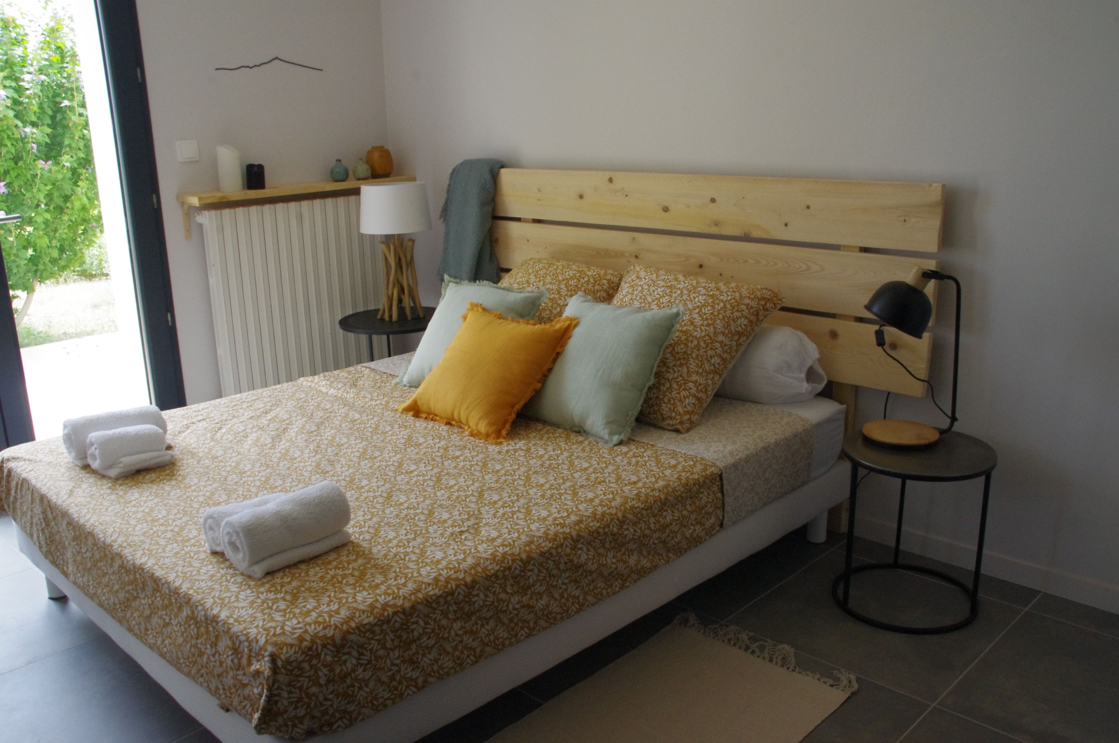 A bedroom with a wooden headboard, a bed with a patterned bedspread, multiple pillows, a bedside table with a lamp, and a view of greenery outside.