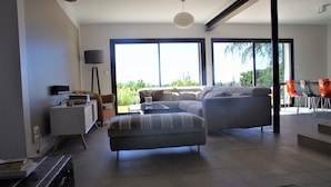 Een moderne woonkamer met een bank, een poef en een salontafel. Er staat een vloerlamp en een bijzettafeltje met een lamp. Grote ramen bieden uitzicht op het omliggende landschap.