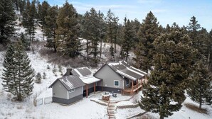 A two-story cabin with a deck, surrounded by snow-covered ground and dense forest. - Colt Mountain Lodge. (Bozeman)