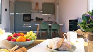 Une table de petit-déjeuner avec un saladier de fruits, des croissants et une tasse de café, dans une cuisine moderne aux armoires vertes et dotée d’un luminaire suspendu.