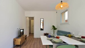Un comedor moderno con una mesa blanca dispuesta para cuatro, rodeada de sillas verdes. Se ve un mueble de televisión de madera a la izquierda, y un espejo grande en la pared.