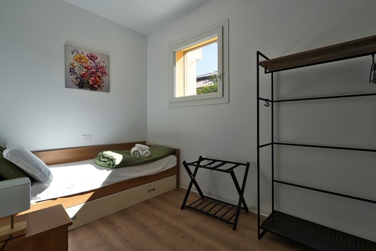 Ein kleiner, heller Raum mit einem Einzelbett, einem Fenster, einem Wandregal und einem Holztisch.