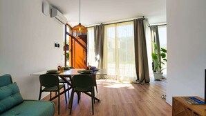 Una sala de estar moderna con comedor, piso de madera, un sofá, una mesa con sillas, una lámpara colgante y un ventanal con cortinas.