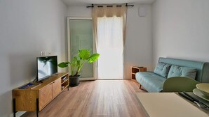 A modern living room with a wooden floor, a television on a wooden cabinet, a green plant, a blue sofa, and a sliding glass door with a beige curtain. - Garden Terrace Apartment w/ A/C & Parking (San Stino di Livenza)