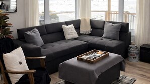 Una sala de estar moderna con un sofá seccional gris oscuro, un sillón mecedor de madera y un pequeño otoman. Se ve un ventilador de techo con lámpara sobre el sofá. Amplias ventanas ofrecen vistas de un paisaje nevado.
