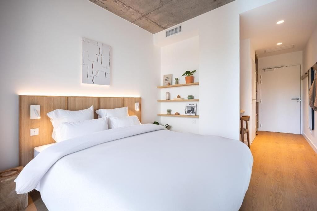 A large bed with a wooden headboard, white bedding, and a map artwork above it. There are shelves with decorative items and a door leading to another room.