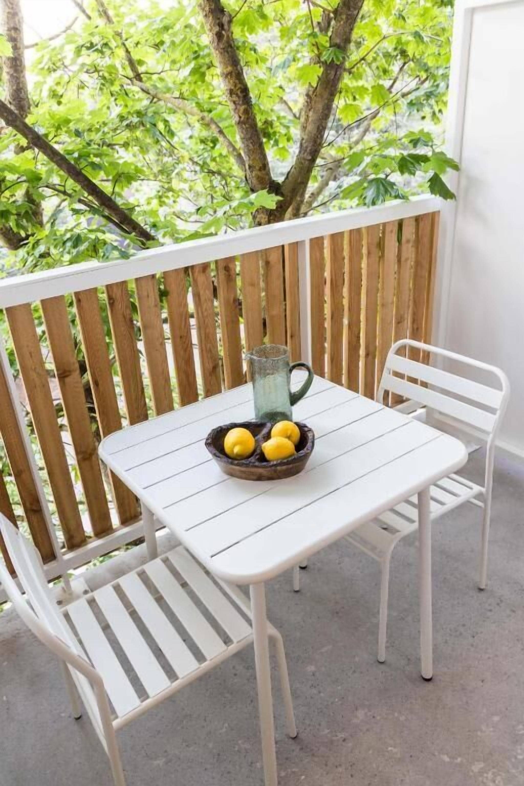 A white table with a bowl of lemons and a pitcher on it, set on a balcony with a wooden railing and trees in the background.