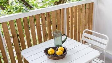 A white table with a bowl of lemons and a pitcher on it, set on a balcony with a wooden railing and trees in the background.