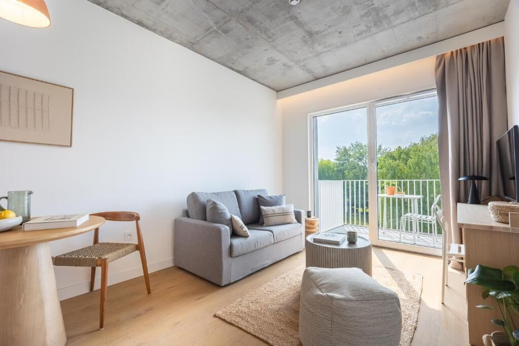 A modern living room with a grey sofa, a wooden dining table, and a small round coffee table. There is a balcony with a view of greenery.