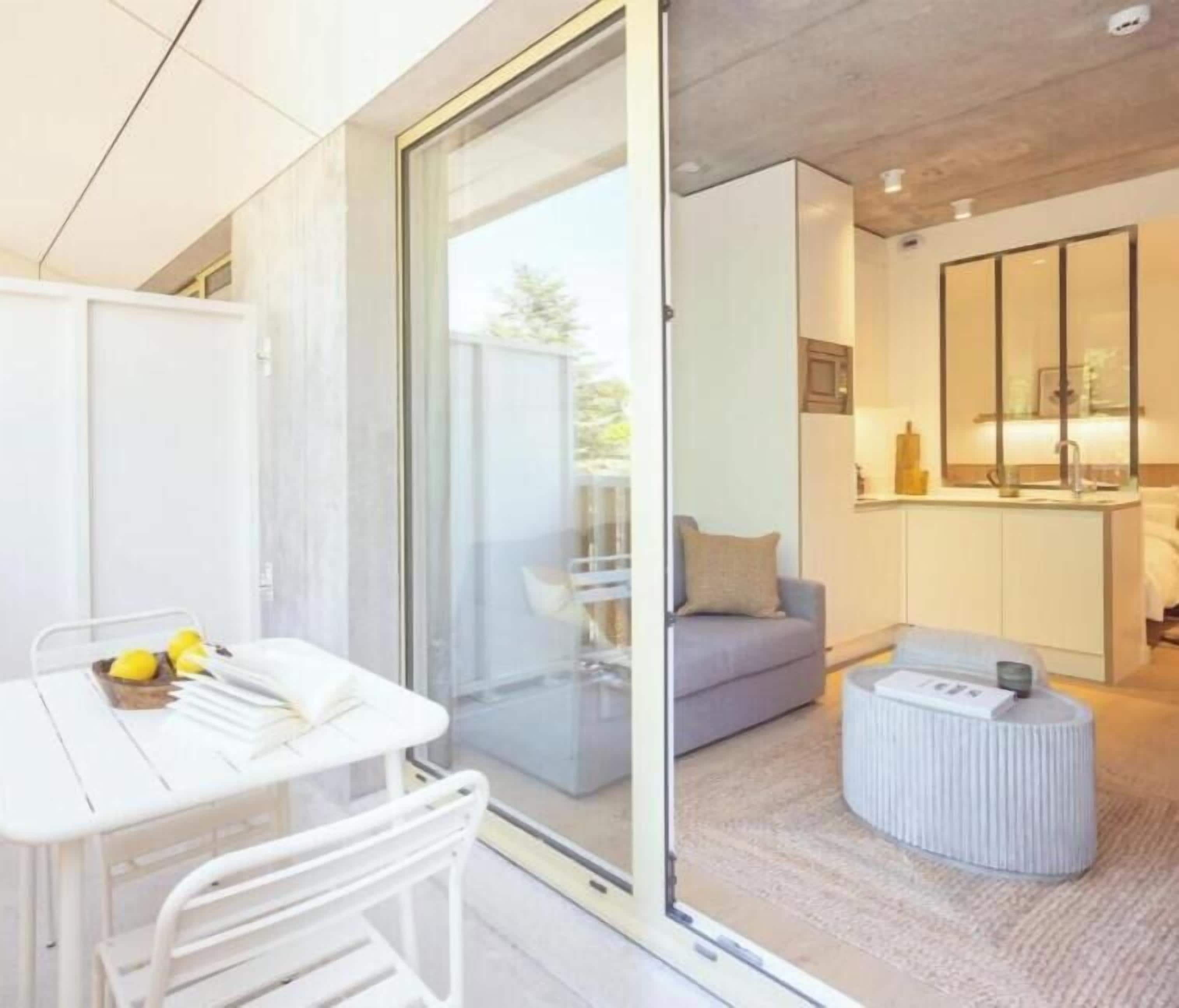 A modern living room with a grey sofa, a white coffee table, and a round ottoman. There is a sliding glass door leading to a balcony with outdoor furniture.
