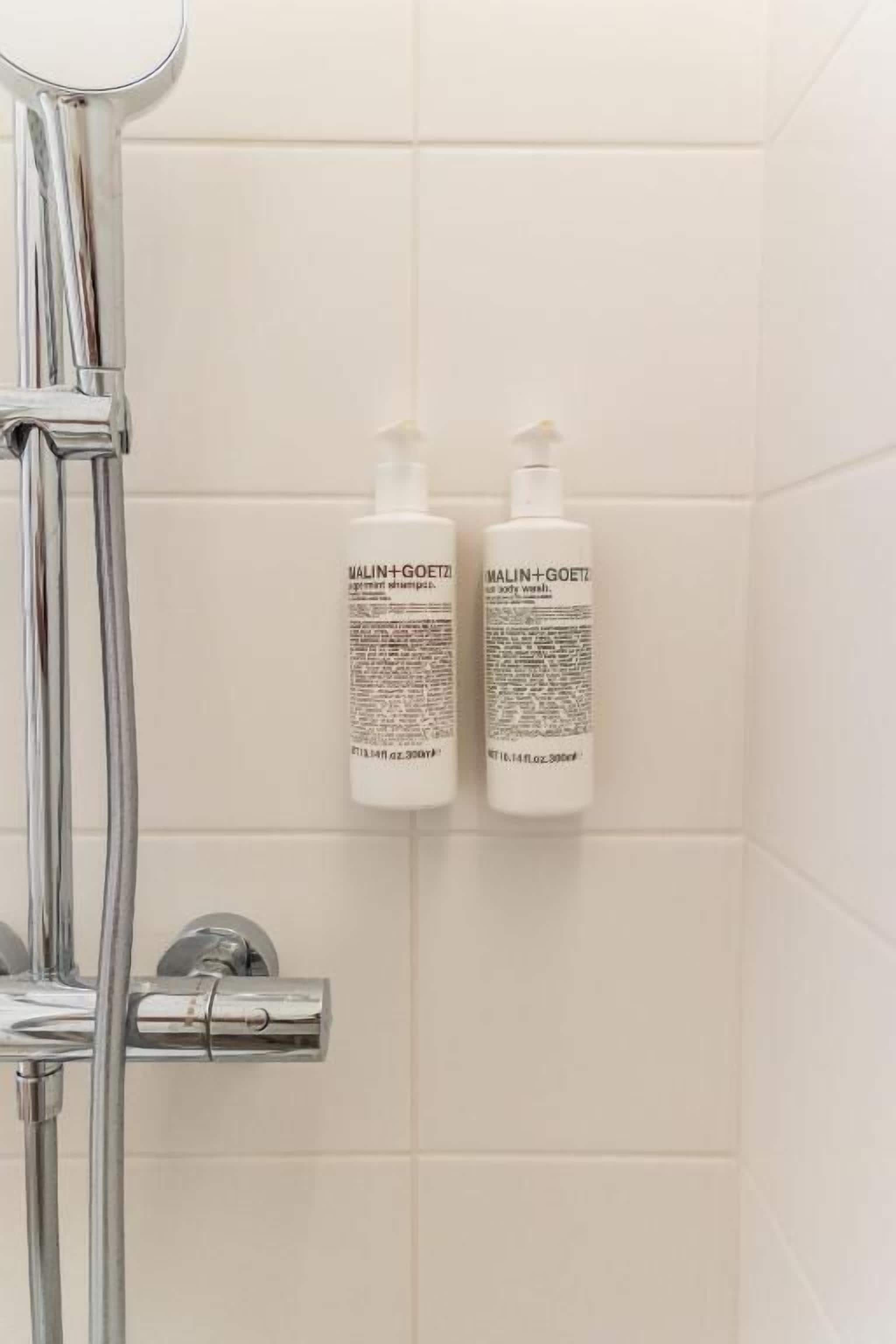 Baño con azulejos blancos, cabezal de ducha cromado y dos botellas de jabón corporal MALIN+GOETZ en la pared.