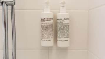 Baño con azulejos blancos, cabezal de ducha cromado y dos botellas de jabón corporal MALIN+GOETZ en la pared.