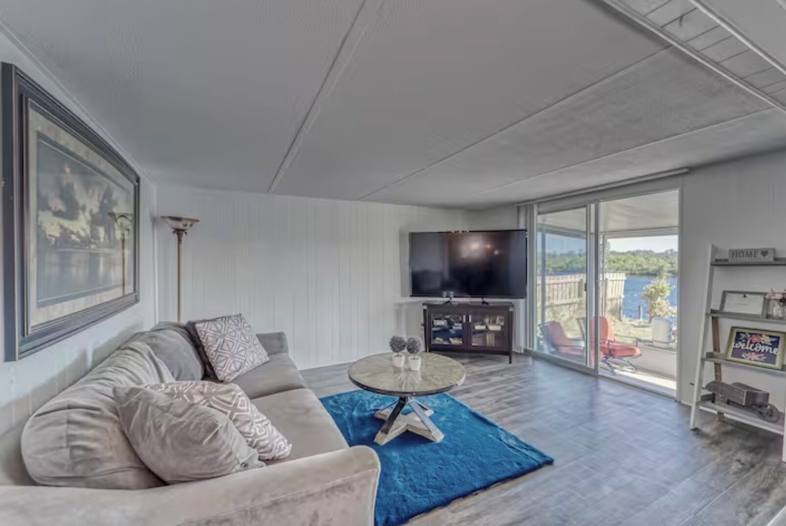A modern living room with a sofa, a coffee table, a flat-screen TV, and a balcony with a view of the outdoors.