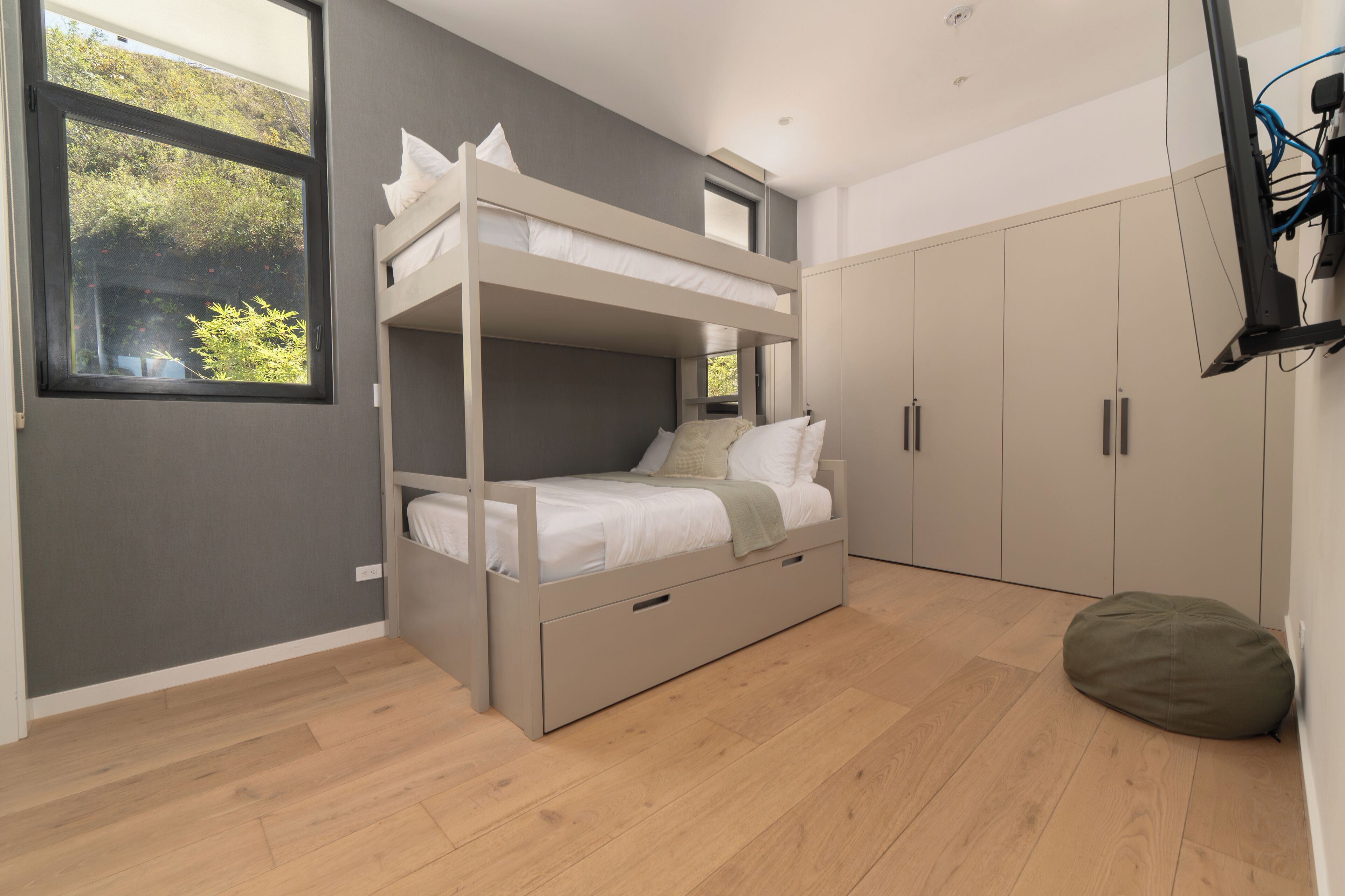 Una cama litera con cajones para guardar objetos, un armario y un sillón orejero en una habitación con suelo de madera y una ventana.