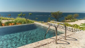 Une piscine aux eaux claires, bordée de pierres et dotée d’un escalier en métal, d’où l’on peut admirer un plan d’eau et des montagnes lointaines.