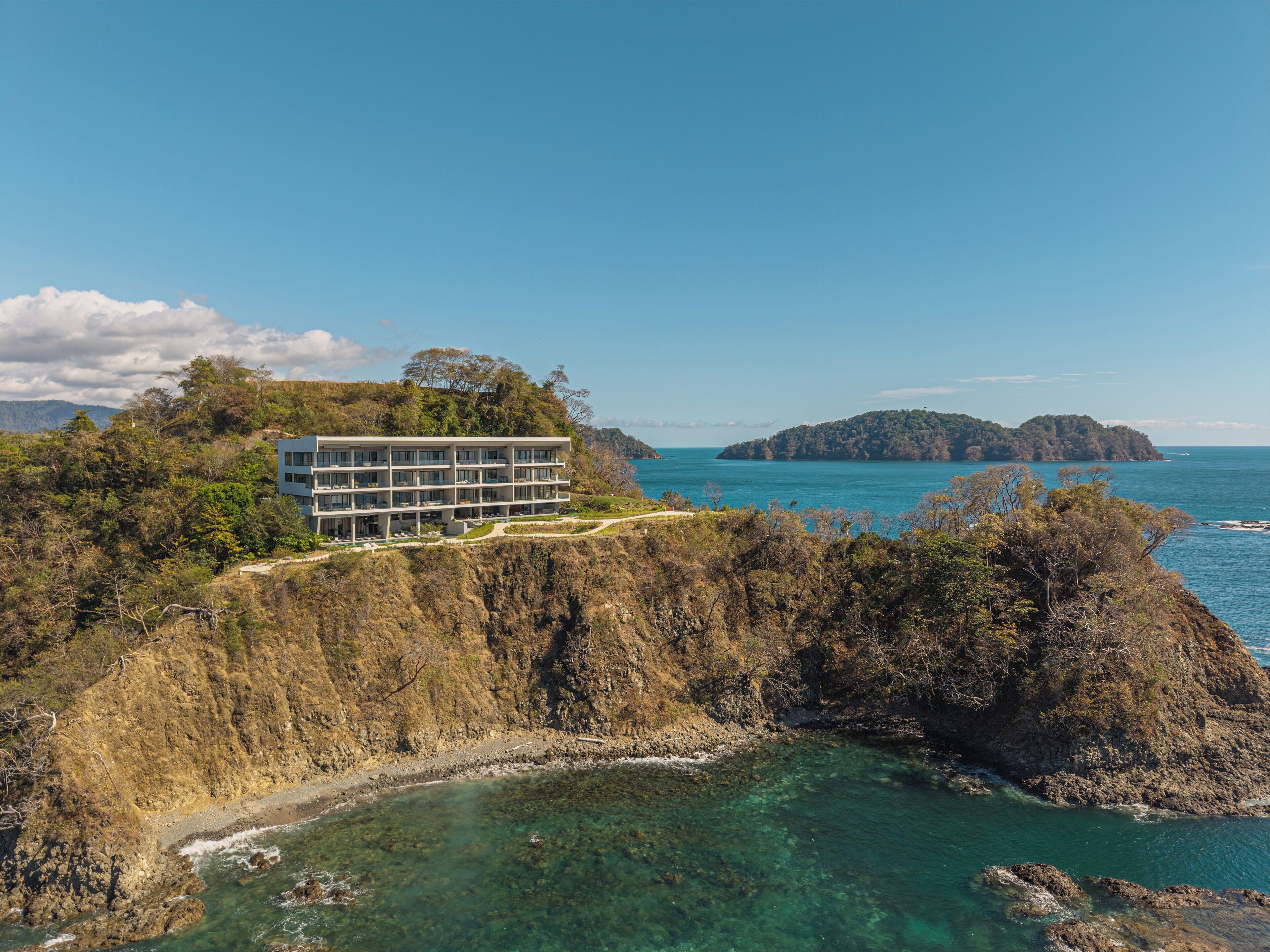 A modern hotel with multiple floors, situated on a cliff overlooking a clear blue sea and a small island.